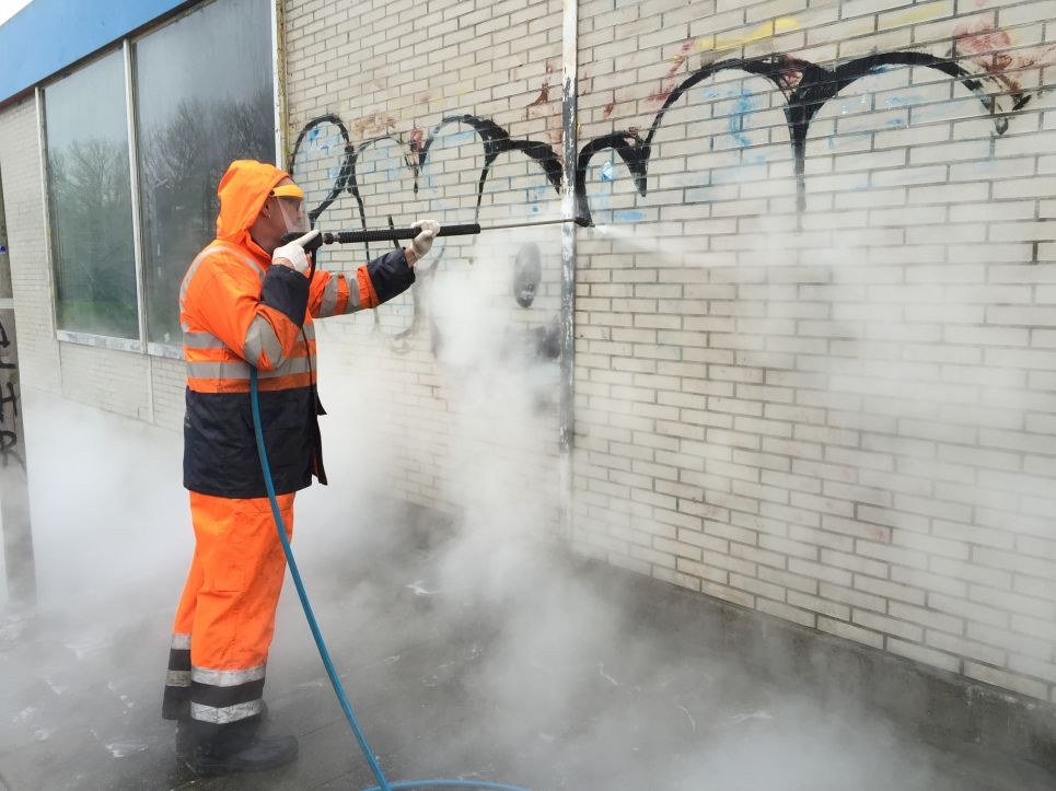Medewerker in oranje werkkleding verwijdert graffiti van een bakstenen muur met een hogedrukspuit